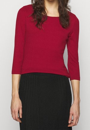 Femme portant un haut rouge côtelé ajusté à manches longues et une jupe noire côtelée, debout devant un fond clair uni.