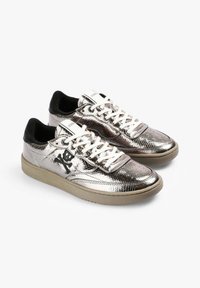 Zapatillas deportivas plateadas metálicas con un acabado texturizado, cordones blancos, acentos negros y un diseño de calavera en el lateral. Suela de goma, forma redondeada.
