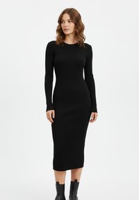 Schwarzes geripptes Midikleid aus Strick mit langen Ärmeln, figurbetonter Silhouette, rundem Ausschnitt und seitlichen Schlitzen, kombiniert mit Stiefeletten.