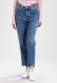 Högmidjade, raka jeans i mediumblå denim med fransad fåll, kombinerade med en långärmad röd- och vitrandig tröja och vita sneakers.