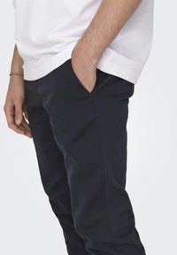 Pantalones azul marino con una textura suave, que presentan bolsillos laterales y un ajuste relajado. Se llevan con una camiseta blanca, parcialmente visible.