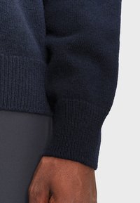 DunkelblauerStrickpulloverbund mit geripptem Muster und glatter Oberfläche, der einen Teil eines dunkelgrauen Kleidungsstücks darunter zeigt.