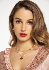 Una donna con capelli castani ondulati indossa un top rosa con volant e collane d'oro a strati. Ha le labbra rosse bold e occhi marroni caldi.
