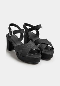 Sandalias de plataforma negras con una textura brillante. Straps cruzados y detalle de hebilla en el tobillo. Tacón cuadrado y grueso. Diseño de punta cerrada.