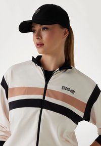 Ženska jakna na patent u bijeloj boji s ružičastim i crnim prugama. Ima crni ovratnik i logo "GUESS 1981". Nosena s crnom kapom.