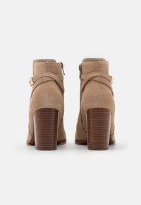 Botas de ante hasta el tobillo en beige claro con punta redondeada, cierre de cremallera y una correa decorativa con una hebilla dorada. Tacón de bloque de madera marrón.