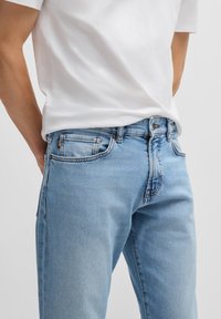 Una persona que lleva una camiseta blanca se encuentra con las manos en las caderas, mostrando unos vaqueros de mezclilla azul claro contra un fondo neutral.
