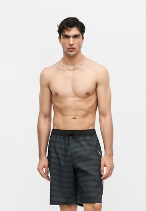 Άνδρας χωρίς πουκάμισο με σκούρα μαλλιά, φορώντας κολιέ με χάντρες και μαύρο σορτς με σχέδια και κορδόνι, μπροστά σε απλό φωτεινό φόντο.