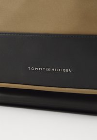Borsa in tela nera e tan con una base in pelle liscia, decorata con un logo argentato che recita "TOMMY HILFIGER". Linee pulite e design moderno.