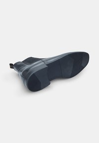 Botín de cuero negro con paneles laterales texturizados, suela de goma plana y tira de arrastre. La parte inferior cuenta con un dibujo sutil para mayor agarre.