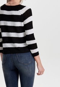 Femme portant un pull en tricot à rayures noires et blanches et un jean bleu, vue de dos sur un fond clair uni.