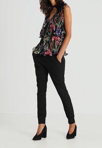 Blusa senza maniche a fiori con volant, caratterizzata da motivi colorati su sfondo nero. Abbinata a pantaloni neri a sigaretta e tacchi.