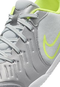 Zapatilla deportiva gris con parte superior de cuero sintético, lengüeta de malla y acentos en amarillo vibrante. Presenta un lado texturizado y suela blanca.