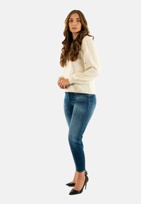 Suéter color marfil con un acabado texturizado, combinado con jeans ajustados azules. El modelo lleva tacones negros de punta, mostrando un cabello liso y un fondo neutro.