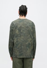 Vyras su pintomis plaukais, dėvintis ilgomis rankovėmis žalią kamufliažinę maršką ir alyvuogių žalumo kelnes, stovintis priešais paprastą baltą foną.