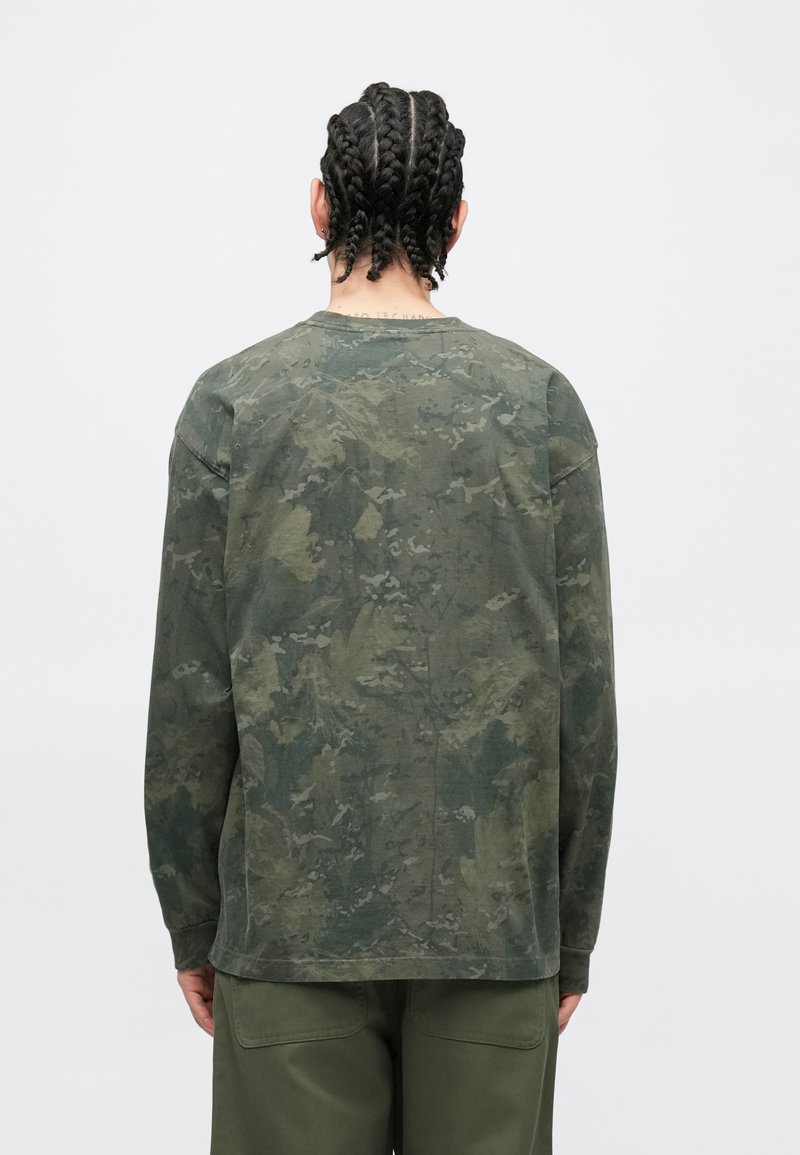 Man with braided hair wearing a long-sleeve green camo shirt and olive green pants, standing facing a plain white background.