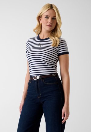 Svetlolaset ženska, oblečena v belo in mornarsko modro črtasto majico s kratkimi rokavi z majhnim logotipom ter temno modre kavbojke z visokim pasom in rjavim pasom.