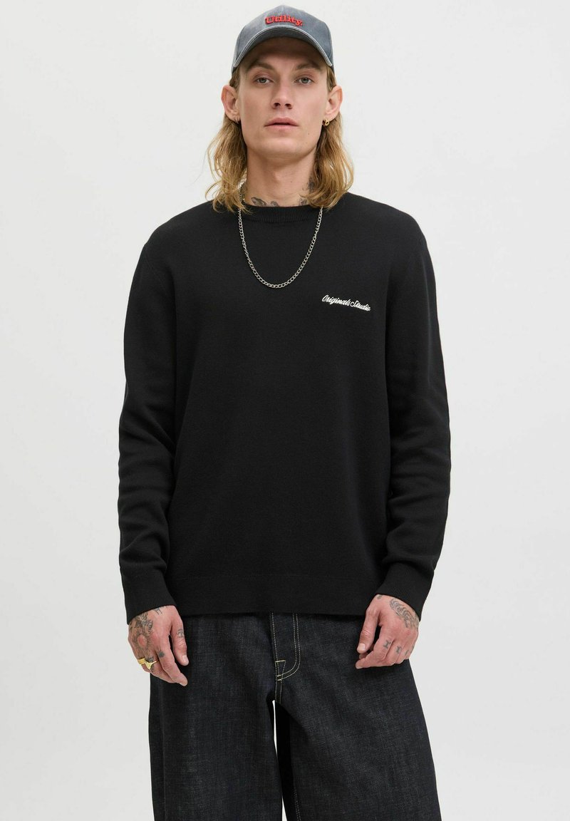 Black sweater with long sleeves and a crew neckline, featuring a small white embroidered logo on the chest. Worn with dark jeans.