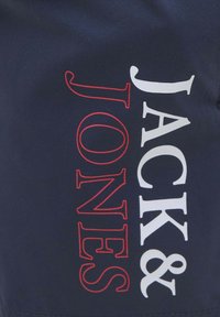 Tela oscura con "JACK & JONES" impreso, "JACK &" en blanco y "JONES" en letras rojas contorneadas, dispuestas verticalmente.