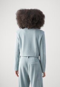 Svetle modrý strukturovaný blazer s kratším střihem, který má žebrovanou látku a dlouhé rukávy. Detail zadní kapsy na odpovídajících kalhotách.