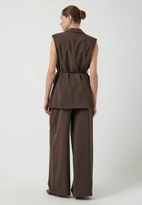 Bruine maatwerk gilet met gestructureerde schouders, een tailleriem en bijpassende wijde broek, gemaakt van textuurstof. Minimalistisch ontwerp.