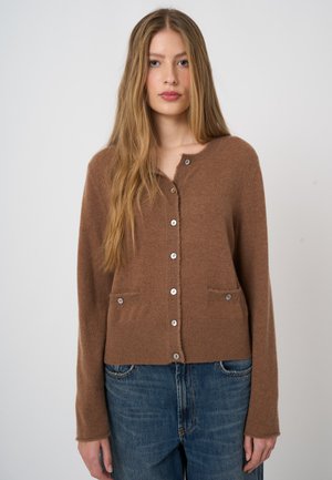 Jeune femme aux longs cheveux blonds porte un cardigan marron boutonné et un jean bleu sur un fond clair uni.
