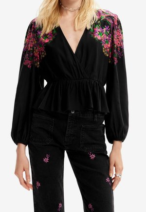 Femme portant un chemisier noir avec une broderie florale rose et violette sur les épaules et un pantalon noir avec une petite broderie de fleur rose.