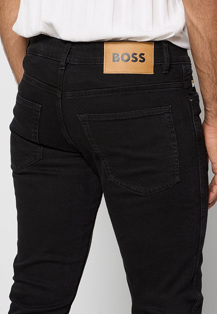 Jean en denim noir avec une taille moyenne, doté de deux poches arrière et d'un logo en cuir brun contrastant sur la ceinture.