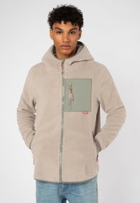 Fleece-Jacke in Hellgrau mit weicher Textur. Verfügt über eine Brusttasche vorne in Grün und orangefarbene Reißverschlussakzente. Mit Kapuze.