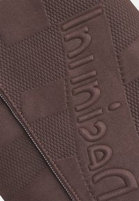 Ruskea, kuvioitu kangas, jossa on kuusikulmainen kuvio ja kohokuvioitu "Huinid"-brändäys. Vetoketju reunan varrella.