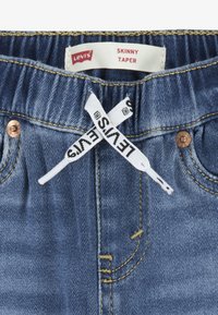 Modre kavbojke iz denima z oznako "Levi's", oznako "Skinny Taper", rumenim šivom in kovinskimi zakovicami. Imajo beli vrvici s črnim besedilom.