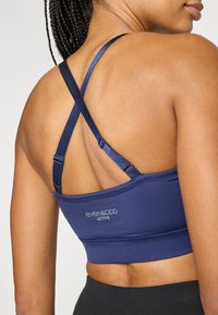 Sportbeha in marineblauw met gekruiste bandjes aan de achterkant, gemaakt van gladde stof met een gebrandmerkt logo en een aansluitend ontwerp. Fijne stiksel details zichtbaar.