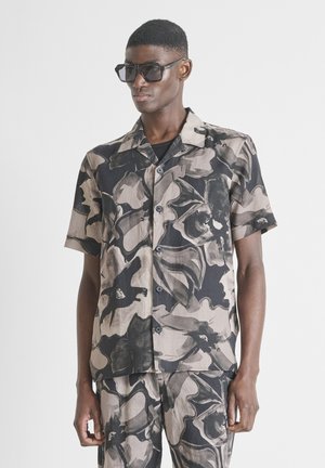 Hombre que lleva una camisa de botón de manga corta con estampado floral abstracto en negro y beige y pantalones a juego con grandes gafas de sol negras.