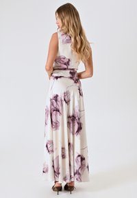 Mouwloze, vloerlange jurk in crème met paarse bloemenpatronen. Gladde stof, getailleerde taille en uitlopende rok. Draag met open schoenen.