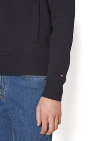 Felpa nera con polsini a coste, tasca laterale e un piccolo logo colorato sul maniche. Abbinata a jeans in denim.