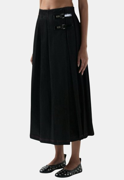Zwarte midirok met plooien en twee glanzende gespsluitingen aan de taille, gedragen met zwarte platte schoenen met zilveren studs.