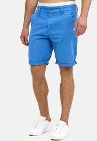 Homme portant un short bleu à ourlet retroussé avec des détails à boutons et des baskets blanches, debout devant un fond clair.
