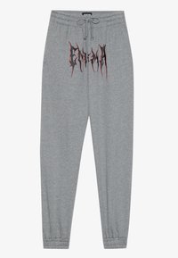 Grå joggebukser laget av myk stoff med tettsittende mansjett, med et svart og rødt grafisk trykk som står "ENIGMA" på framsiden.