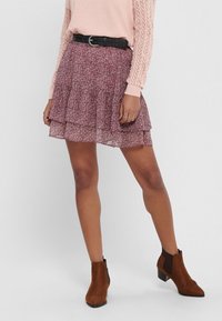 Jupe florale mauve superposée avec un voile transparent, en dessous du genou. Associée à un pull rose clair et des bottines brunes.
