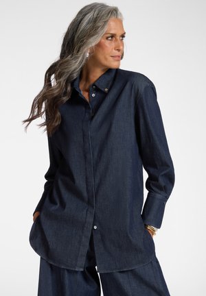 Vrouw met lang grijs haar draagt een donkerblauw spijkerblouse en broek, kijkt naar haar rechterkant met handen in de zakken tegen een witte achtergrond.