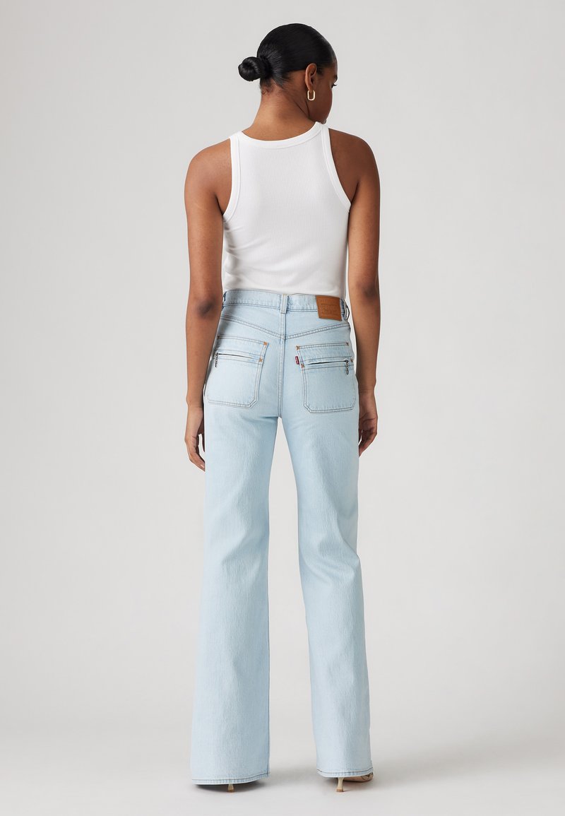 Jean taille haute bleu clair avec des jambes évasées, comportant une étiquette en cuir marron à l'arrière et deux poches zippées à l'arrière.