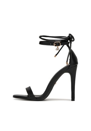 Czarna sandał na wysokim obcasie z cienkim paskiem wokół kostki, wiązaniem, odkrytymi palcami i minimalistycznym paskiem z przodu, z małą złotą klamrą i detalem w postaci zawieszki.