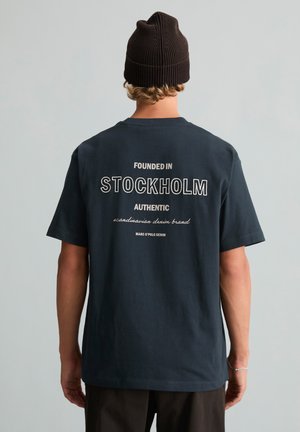 Personne portant un bonnet noir et un t-shirt foncé avec un texte blanc "Founded in Stockholm Authentic Scandinavian denim brand" dans le dos.