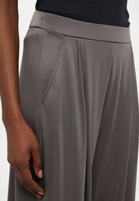 Pantaloni grigi con una texture liscia, caratterizzati da una vita aderente e plissettature laterali. Mano poggiata sulla zona della tasca, che mette in mostra il drappeggio del tessuto.