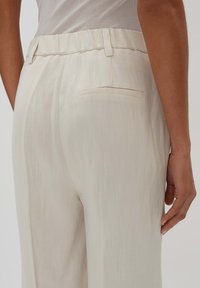 Beige, lichte broek met een gestructureerde stof, een elastische tailleband en een enkele achterzak. Losse pasvorm met rechte pijpen.