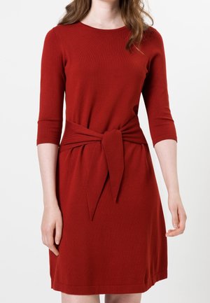 Woman wearing a red knee-length dress with three-quarter sleeves and a fabric belt tied at the waist against a white background.