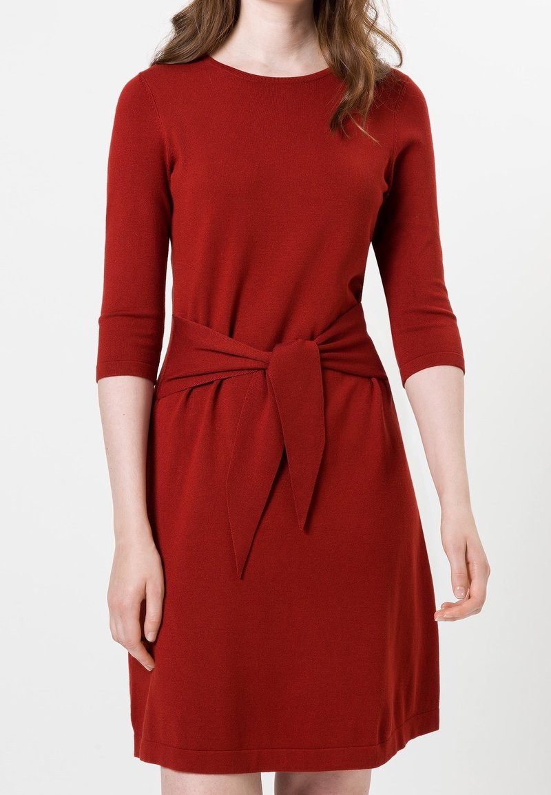 Femme vêtue d'une robe rouge longueur genou avec des manches trois-quarts et une ceinture en tissu nouée à la taille sur fond blanc.
