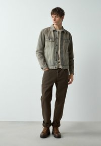 Pelēks džinsu jaciņa ar divām krūšu kabatām, valkāta virs krēmkrāsas džempera, saskaņota ar tumši brūnām biksēm un brūnām šņorzābakiem.