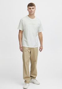 Lys grønn kortermet t-skjorte med "HVORFOR HASSE?" i gult tekst; kombinert med beige bukser og hvite joggesko.