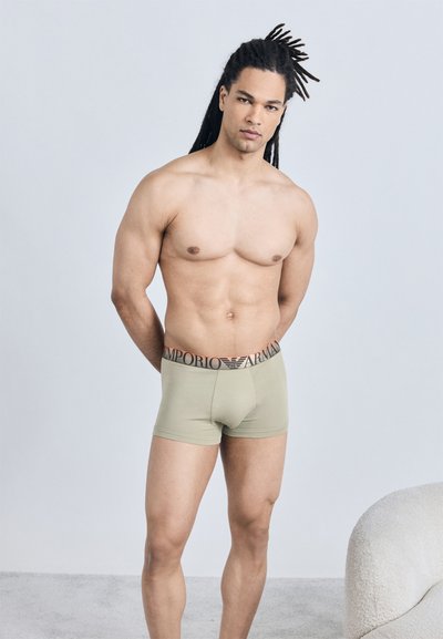 Άνδρας χωρίς πουκάμισο με dreadlocks στέκεται, φοράει μπεζ boxer briefs Emporio Armani μπροστά σε απλό φωτεινό φόντο.